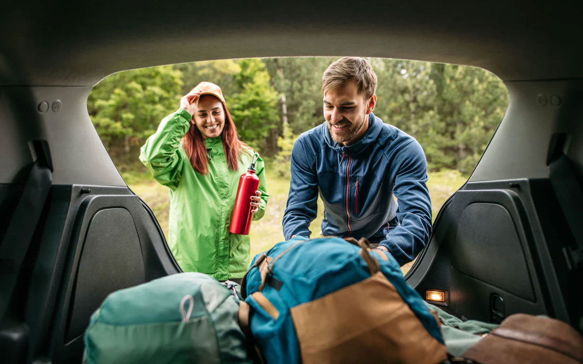 Una ragazza e un ragazzo tirano fuori dal bagagliaio di una macchina i loro zaini da trekking