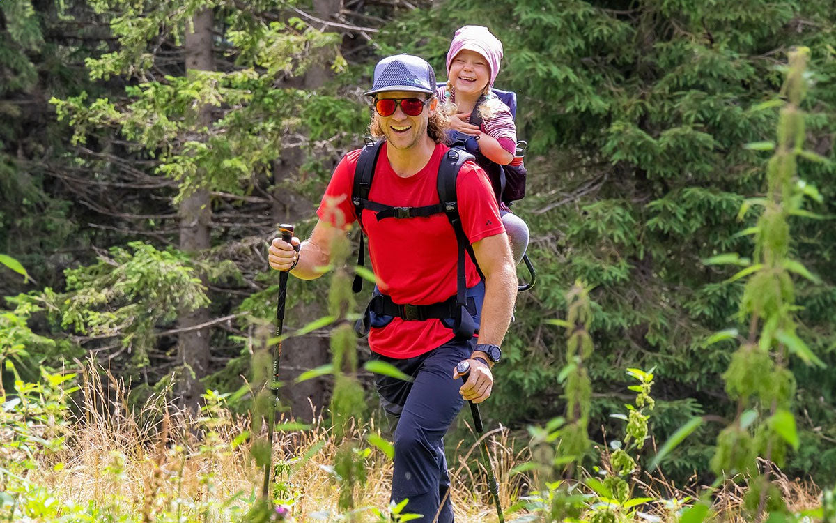 Un ragazzo con una bambina in spalla cammina utilizzando bastoncini da trekking Leki
